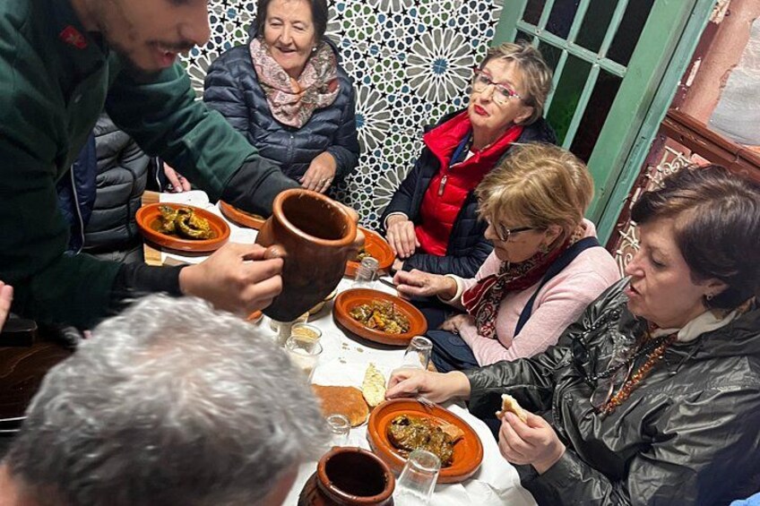 Een groep mensen dineert aan een tafel met traditionele Marokkaanse gerechten en een grote terracotta pot. 3