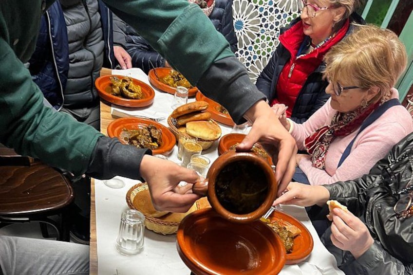 Mensen delen een maaltijd aan een tafel met verschillende gerechten en een grote terracotta pot. 4