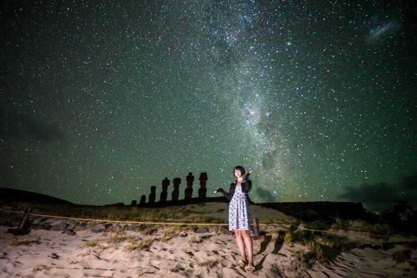 A person standing on a sandy beach with a starry sky above and a row of stone statues in the background. 4