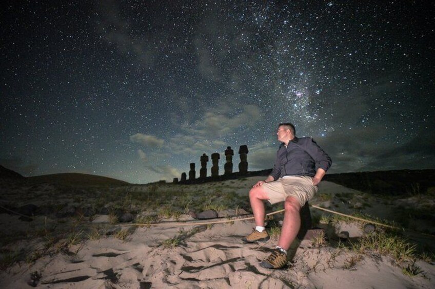 A person sitting on a sandy area under a starry sky with a row of stone statues in the background. 5