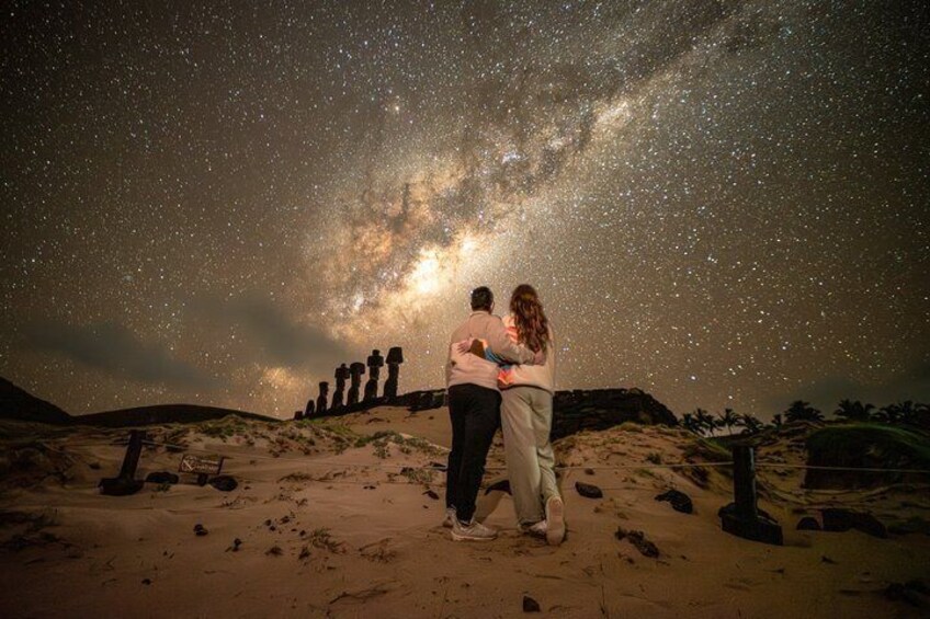 A couple standing on a sandy beach under a starry sky with a Milky Way view, with ancient stone structures in the background. 1
