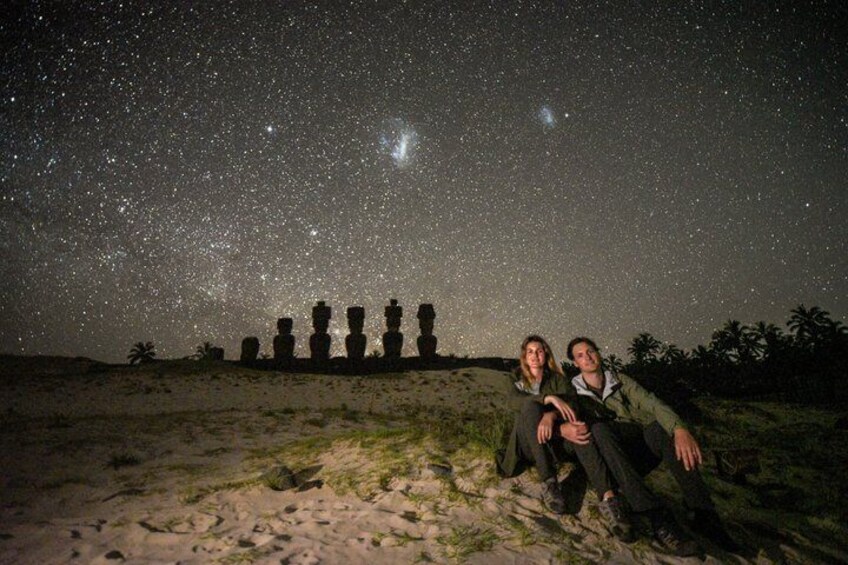 Two people sitting on a sandy beach with a starry night sky above and a row of stone statues in the background. 2
