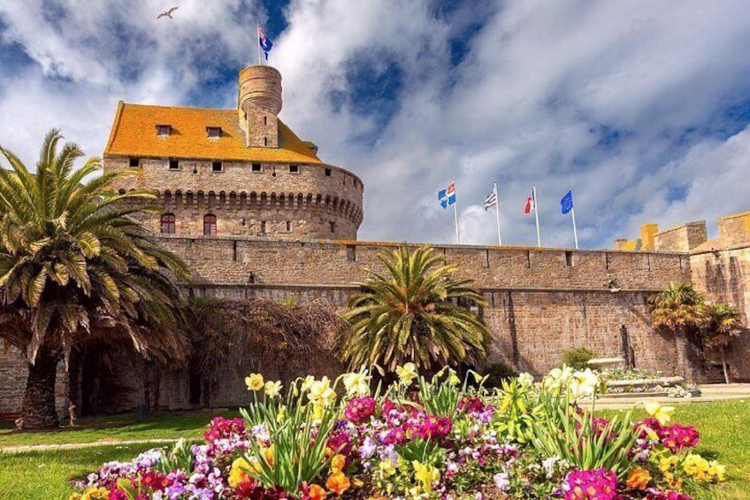 A historic stone fortress with a round tower, palm trees, and colorful flowers in the foreground. 4