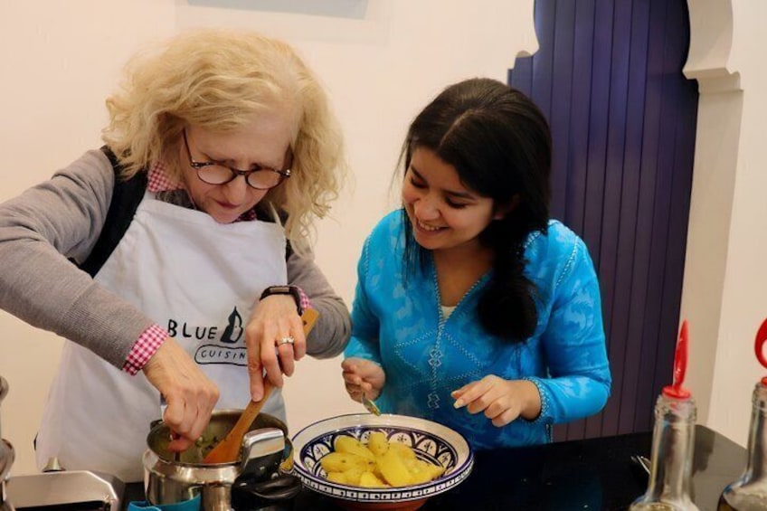 Two people cooking, one wearing a white apron with 'BLUE CUISINE' text, and another in a blue top. 1