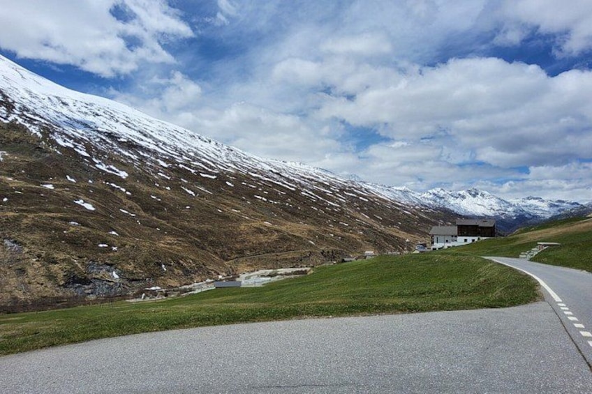 Una carretera sinuosa atraviesa un paisaje montañoso con picos nevados, una zona cubierta de hierba y un edificio a lo lejos. 4