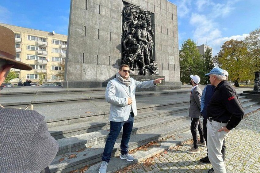 Ein Mann deutet auf ein großes Denkmal mit einem Relief, umgeben von einem gepflasterten Bereich und Gebäuden. 1