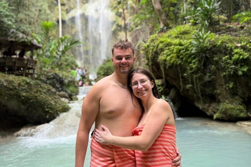 A couple embracing in a lush, green forest with a waterfall in the background. 3