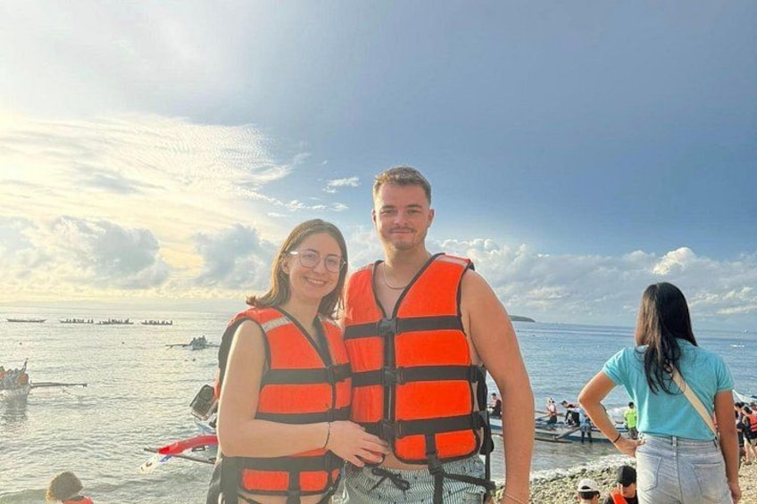 Two people wearing life jackets on a beach with boats in the background. 5