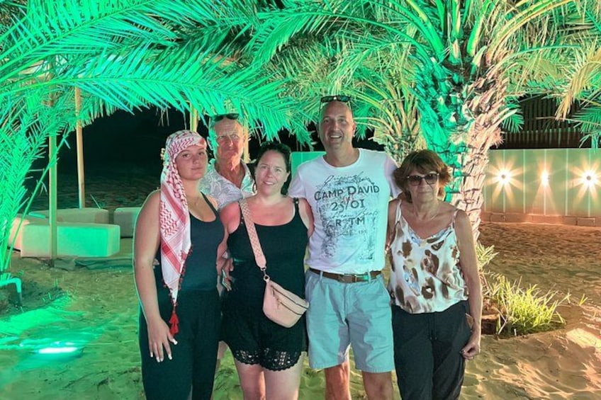 A group of people standing under a palm tree with a beach and a building in the background. 3