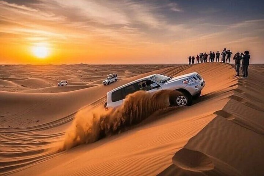 Un SUV bianco che percorre un deserto con dune di sabbia, un tramonto sullo sfondo e un gruppo di persone che osservano di lato. 2