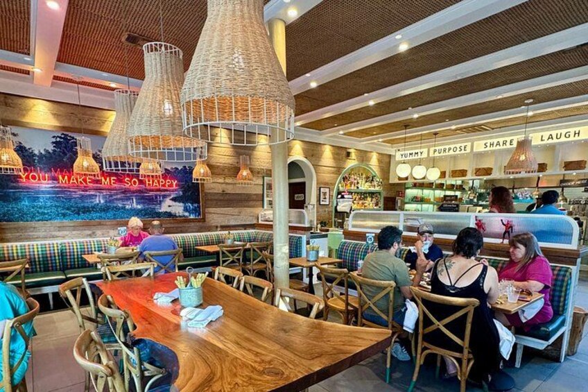 Interior de restaurante con mesas y sillas de madera, lámparas de mimbre colgantes y una pared grande con una cita motivacional. 1