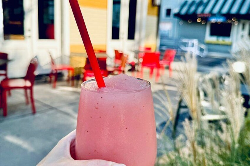 Una bebida rosa con pajita roja en un vaso, sobre un fondo borroso de un café al aire libre con sillas y mesas rojas. 2