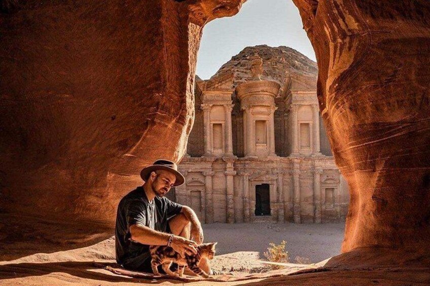 A person sitting in a natural rock formation with a camel in front of a historic building. 1