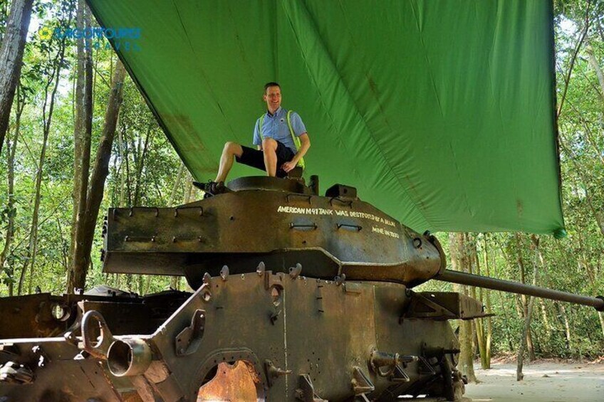 A person sitting on a military tank with a green canopy, surrounded by trees. 2