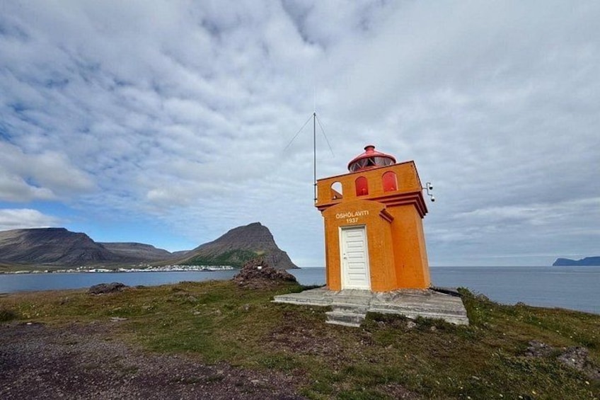 A lighthouse named 'Osholaviti' with a red roof, situated on a grassy hill with a view of the sea and mountains in the background. 2