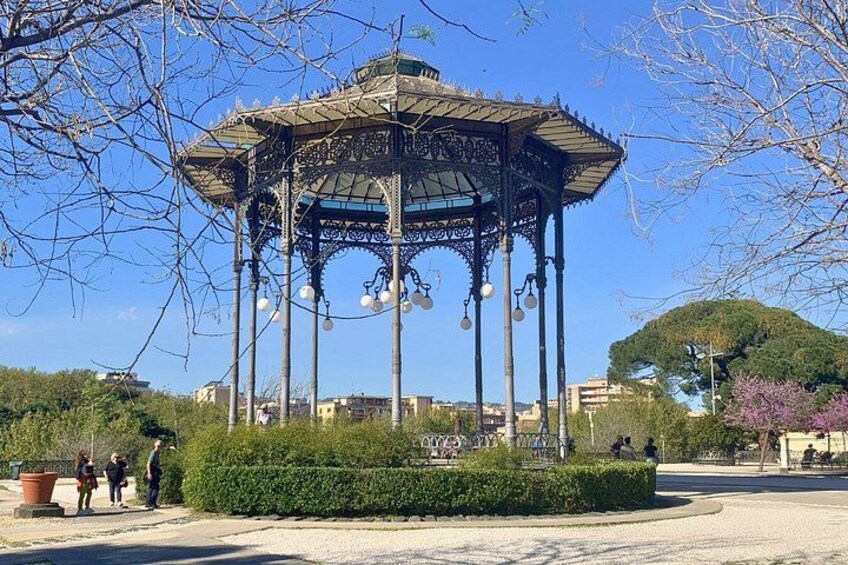 A gazebo with a glass top, surrounded by a well-maintained hedge, in a park with trees and buildings in the background. 4