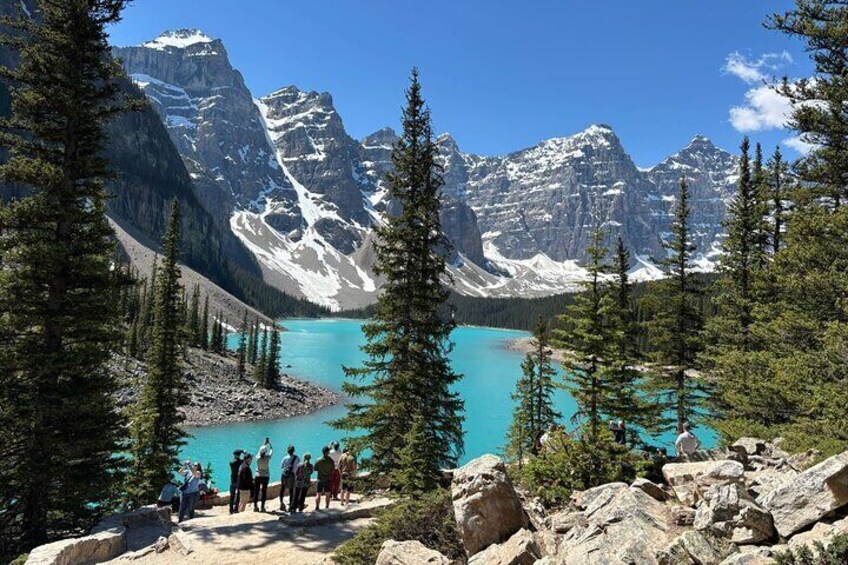 Un gruppo di persone in piedi su una sporgenza rocciosa con vista su un lago turchese e montagne innevate sullo sfondo. 5
