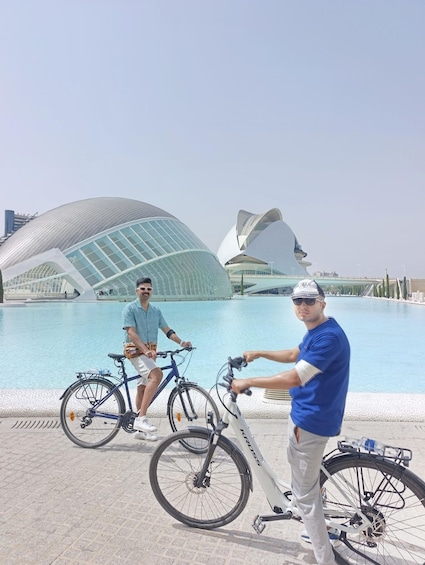 Two people with bicycles near a modern architectural structure by a body of water. 2
