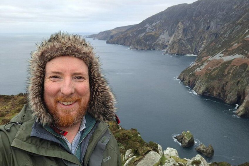 A person in a fur-lined jacket overlooking a coastal landscape with rocky cliffs and a body of water. 4