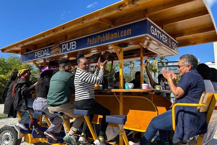 En trækonstruktion med teksten 'Pedal Pub Miami.com', hvor folk sidder og står rundt omkring. 2