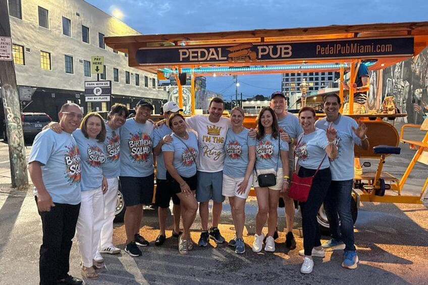 A group of people in front of a Pedal Pub with a sign and a vehicle. 1