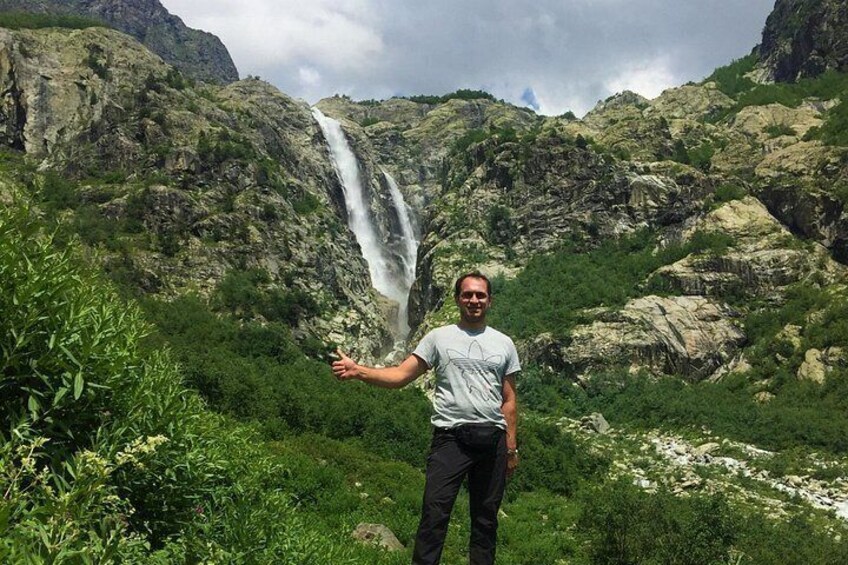 A person standing in front of a waterfall with lush greenery and rocky terrain in the background. 4