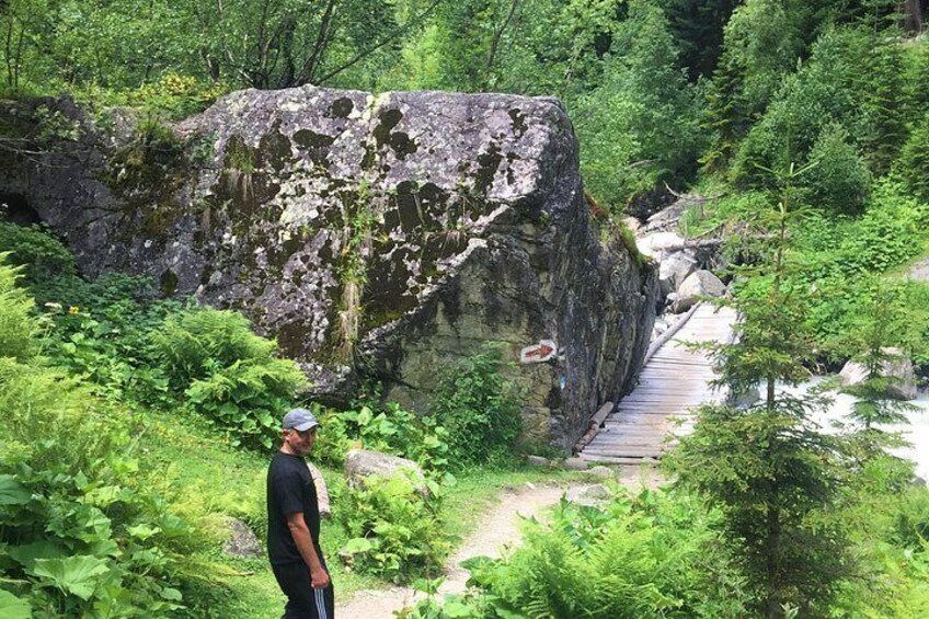 A person standing on a wooden bridge over a river, surrounded by lush greenery and a large rock formation. 2