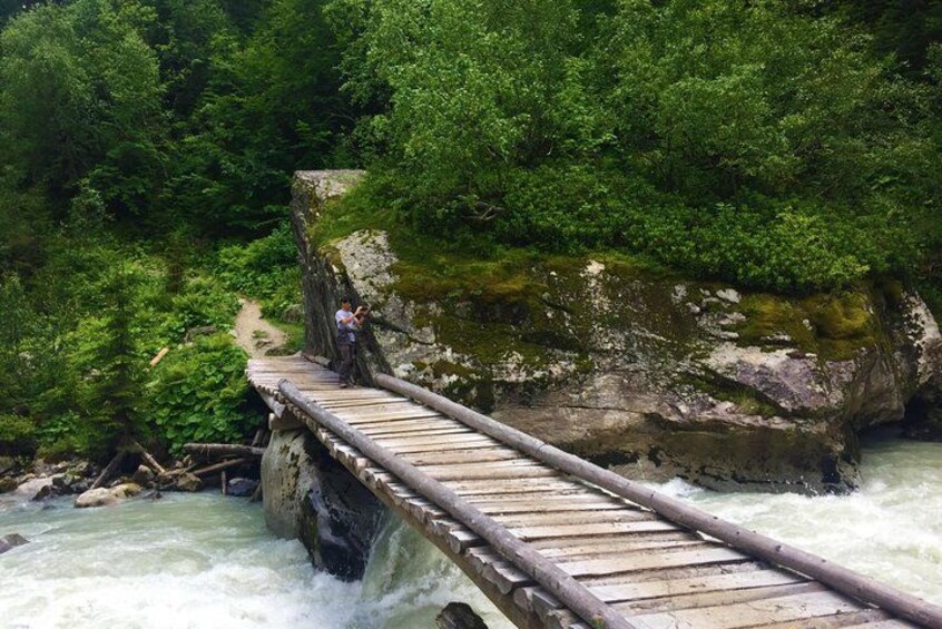 A wooden bridge over a fast-flowing river, surrounded by lush greenery and rocky cliffs. 2