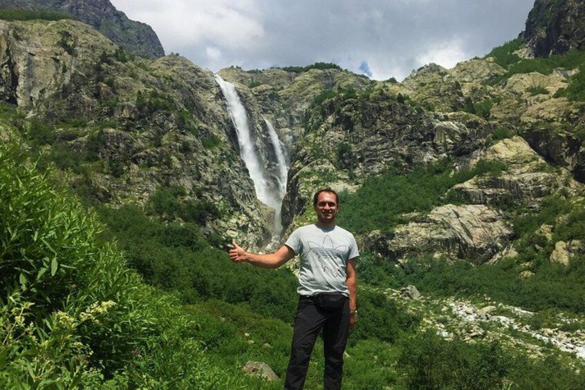 A person standing in front of a waterfall with lush greenery and rocky terrain in the background. 5