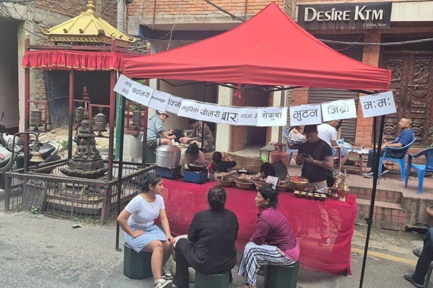 A street food stall with a red canopy, people sitting and eating, and a signboard with text. 3