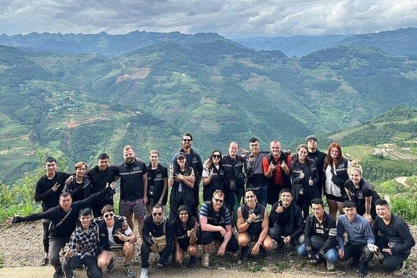 A group of people posing on a mountain ridge with a panoramic view of green valleys and distant hills. 2