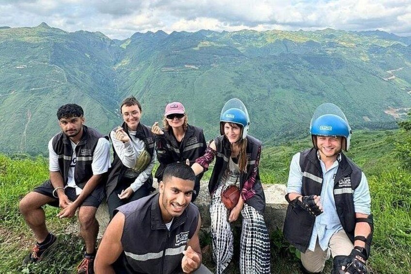 A group of people in outdoor gear sitting on a grassy hill with a mountainous landscape in the background. 3