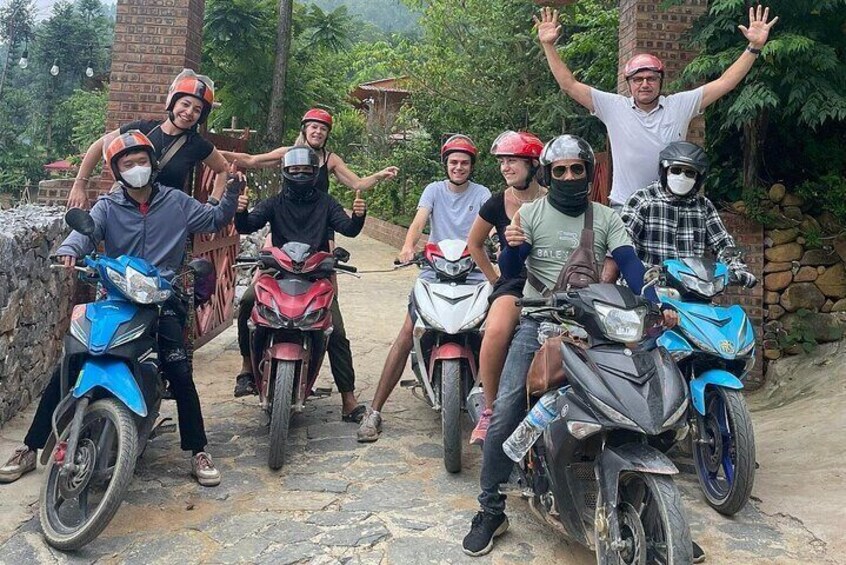 A group of people on motorcycles, some wearing helmets, posing on a stone path with greenery in the background. 1