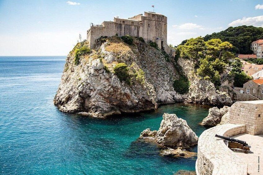 A historic stone fortress on a rocky cliff by the sea, with clear blue waters and a small town in the background. 2