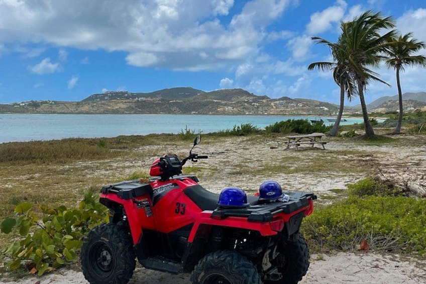 En rød ATV med hjelme bagpå, en sandstrand, palmer og et vandareal med bakker i baggrunden. 5
