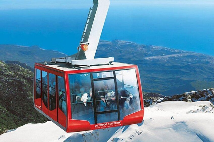 A red cable car with passengers, suspended by a cable, overlooking a mountainous landscape with a body of water in the distance. 1