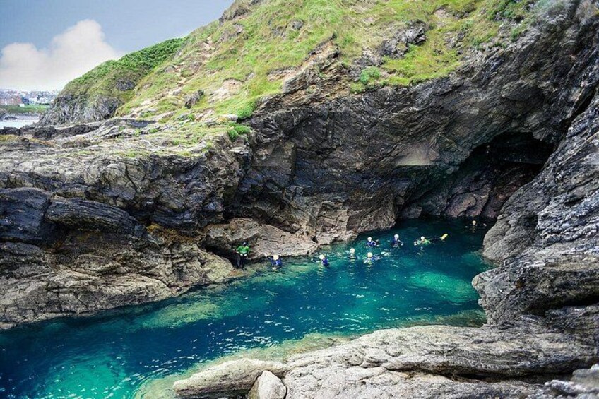 A rocky coastal area with clear blue water, a small cave entrance, and people swimming. 5