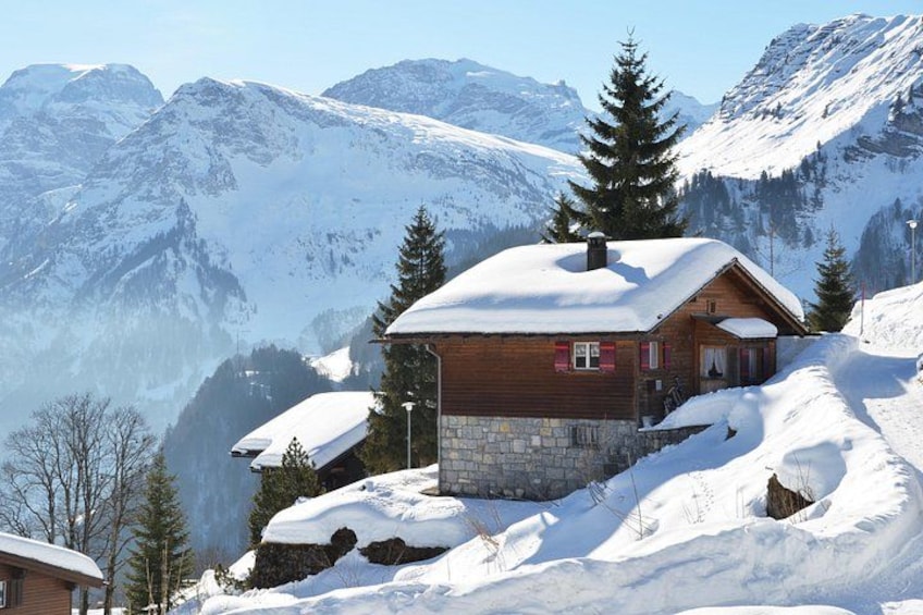 Une cabane en bois, dont le toit est recouvert de neige, entourée de montagnes et d’arbres également couverts de neige. 3