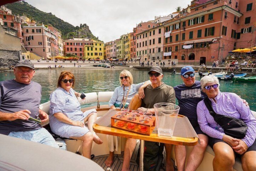 A group of people on a boat with a table of food and drinks, surrounded by colorful buildings and water. 1
