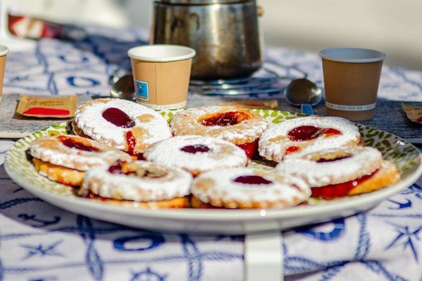 En tallerken med kringler drysset med flormelis, en kaffekop og en metalboks på en mønstret dug. 4