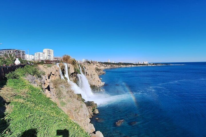 Ein Küstenwasserfall mit klarem, blauem Wasser, einem felsigen Felsen und Gebäuden im Hintergrund. 1