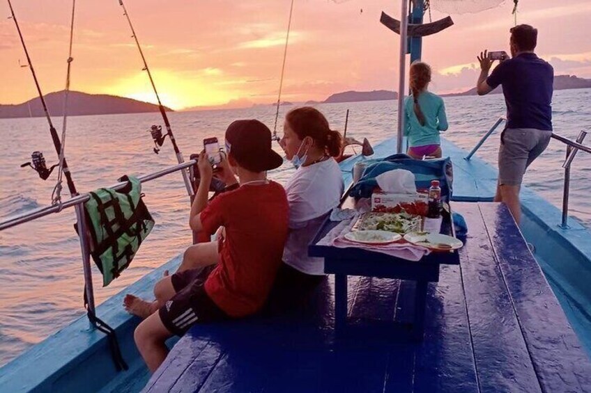 A boat with people sitting and taking photos, a table with food, and a sunset in the background. 5