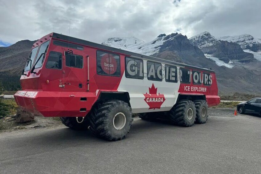 A red and white Glacier Tours ice explorer vehicle with a Canadian flag, parked on a road with mountains in the background. 5