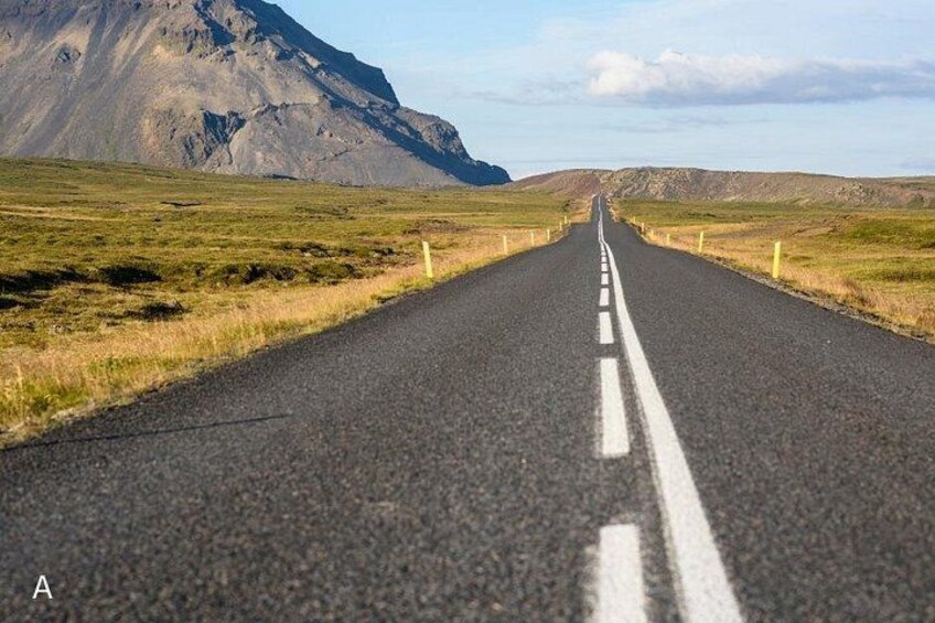 A road with a white center line, flanked by grassy fields and mountains in the distance. 3