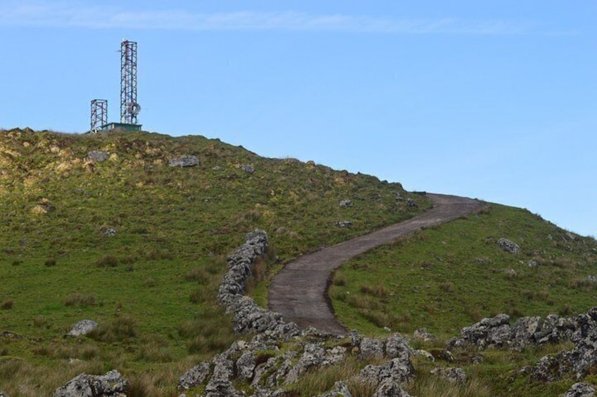 A winding road on a grassy hill with a stone wall and two communication towers in the distance. 5