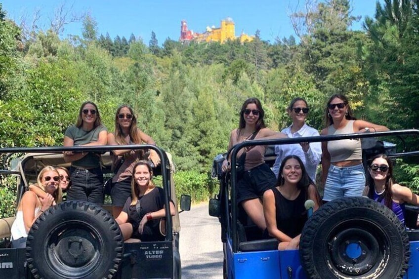 Un groupe de personnes se trouve dans un jeep sur une route bordée de verdure, avec un bâtiment visible au loin en arrière-plan. 1