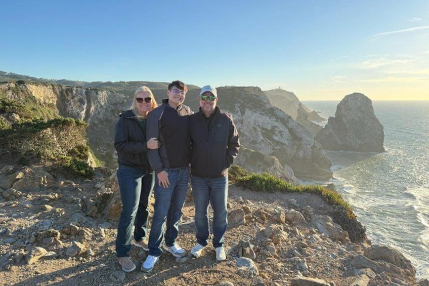 Trois personnes se tiennent debout sur une falaise rocheuse, surplombant l’océan, avec d’autres falaises en arrière-plan. 5