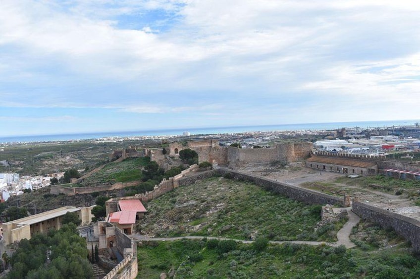A historical fortification with a surrounding wall, a pathway, and a view of the city and sea in the distance. 3