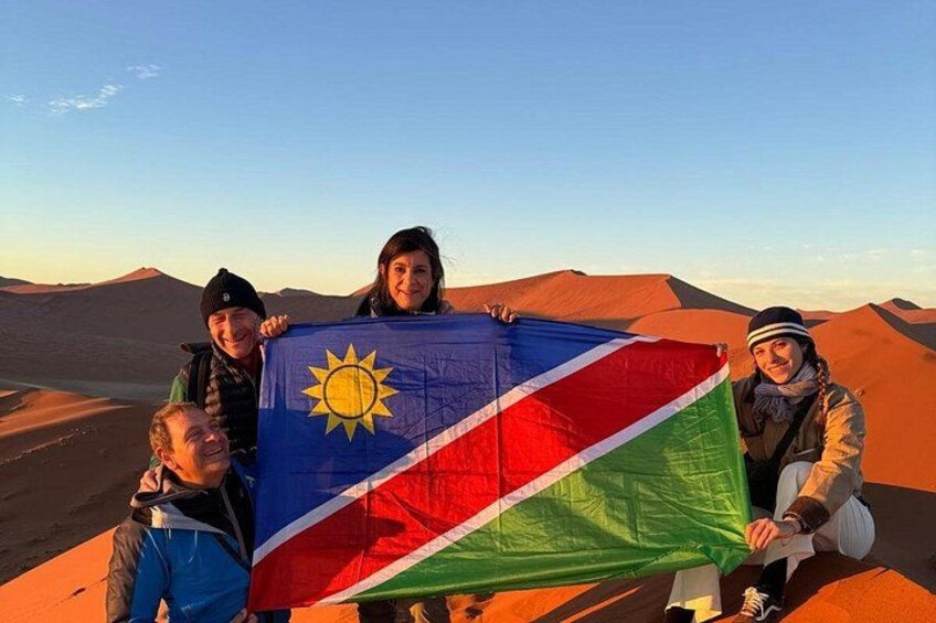 A group of people holding a flag with a sun emblem, standing in a desert landscape with sand dunes. 5