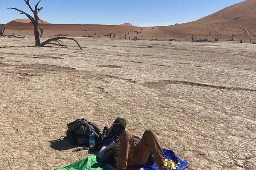 A person lying on a blue mat in a dry, barren landscape with sparse vegetation and a clear sky. 2